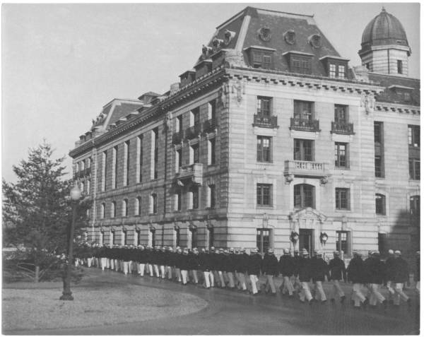 Midshipmen marching, alongside a wing of Bancroft Hall at USNA, in 'white works' uniform wearing 'reefers,' to some event.